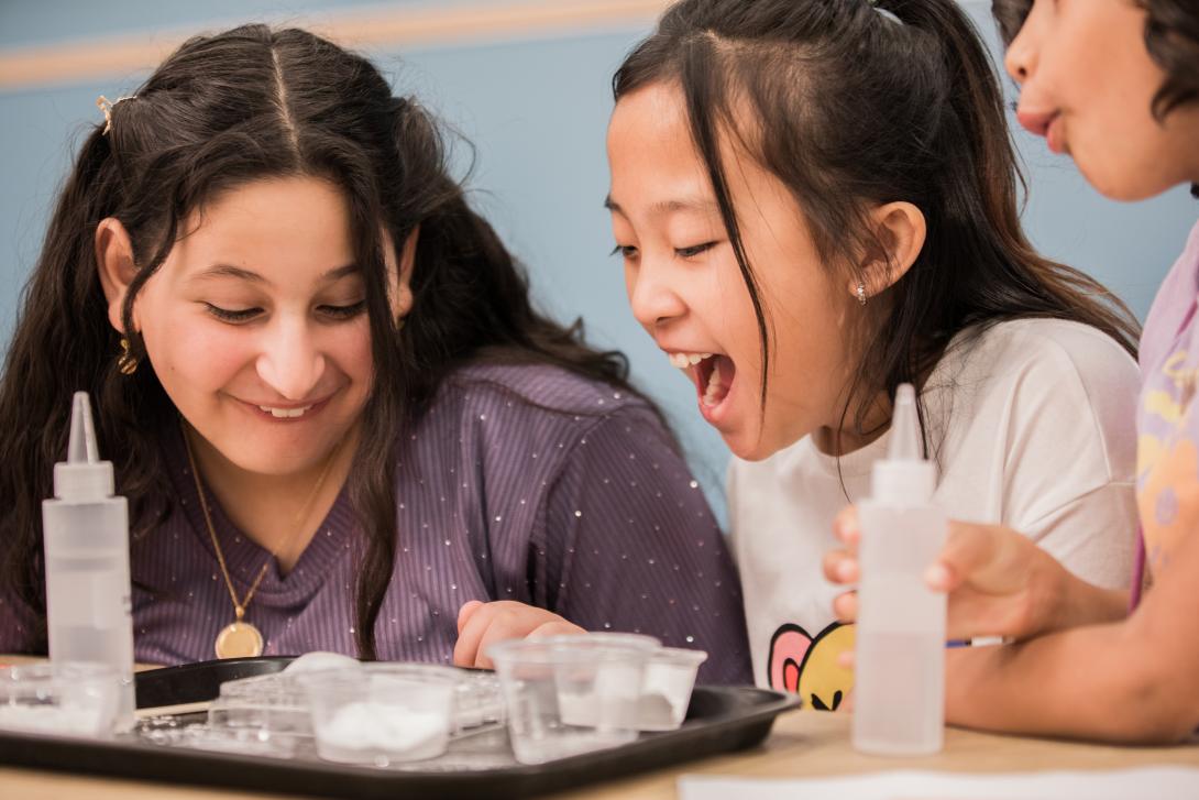 3 young girls smile and exclaim at a chemical reaction in a plastic cup on a brown tray on the table